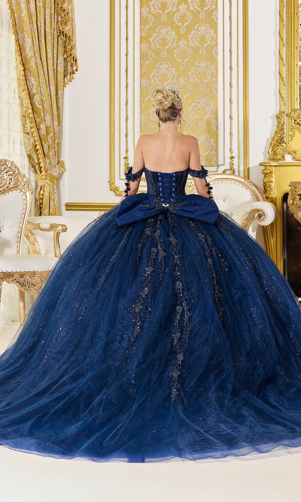 A woman wearing the Quinceanera Dress 5083 By Dancing Queen, an off-the-shoulder navy blue ball gown with a sparkling full skirt, stands facing away in an elegantly gold-accented room.